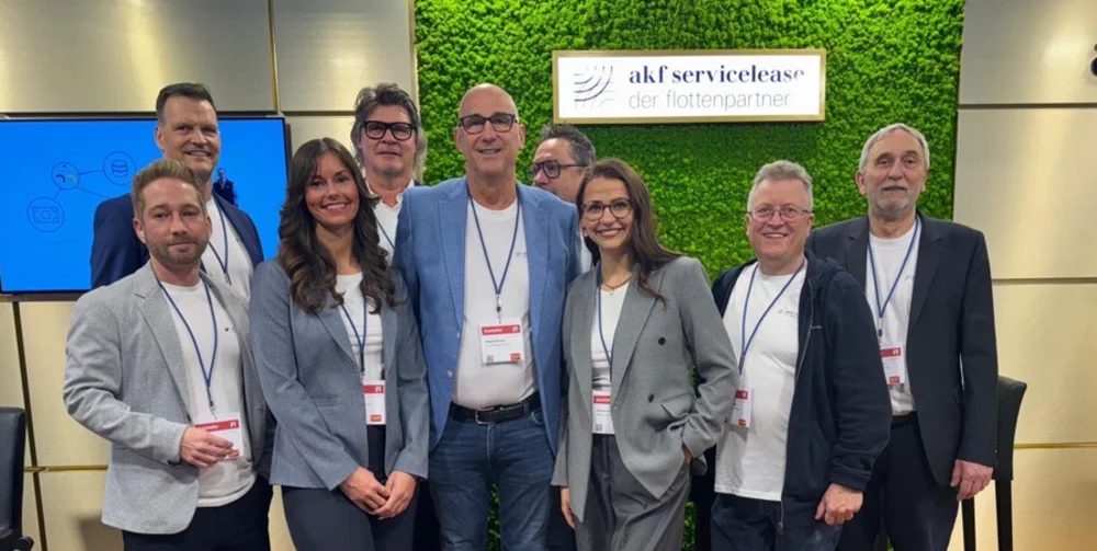 Gruppe von acht Personen mit Namensschildern steht vor grünem Wandpaneel mit Schild akf servicelease der flottenpartner und zwei Bildschirmen