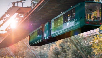 Schwebebahn mit zwei Waggons fährt auf einer Stahlkonstruktion bei Sonnenuntergang über bewaldetes Gelände