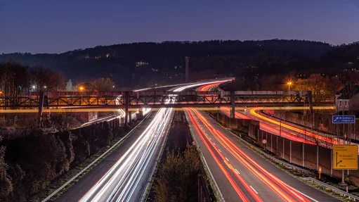 Langzeitbelichtete Aufnahme einer mehrspurigen Autobahn bei Nacht. Die Scheinwerfer und Rücklichter der Fahrzeuge erzeugen lange Lichtspuren, die weisse und rote Linien auf der Fahrbahn bilden. Im Hintergrund sind eine Brücke und bewaldete Hügel zu sehen. Strassenbeleuchtung und Verkehrsschilder sind sichtbar, darunter ein Schild mit Richtungen zu den Städten Solingen und Remscheid.