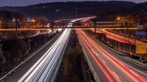 Langzeitbelichtete Aufnahme einer mehrspurigen Autobahn bei Nacht. Die Scheinwerfer und Rücklichter der Fahrzeuge erzeugen lange Lichtspuren, die weisse und rote Linien auf der Fahrbahn bilden. Im Hintergrund sind eine Brücke und bewaldete Hügel zu sehen. Strassenbeleuchtung und Verkehrsschilder sind sichtbar, darunter ein Schild mit Richtungen zu den Städten Solingen und Remscheid.