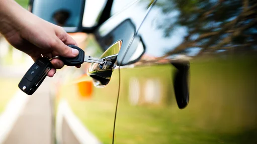 Nahaufnahme einer Hand, die einen Autoschlüssel in das Schloss eines glänzenden, schwarzen Autos steckt. Die Autotür und der Hintergrund spiegeln die Umgebung mit unscharfem Gras und Bäumen wieder.